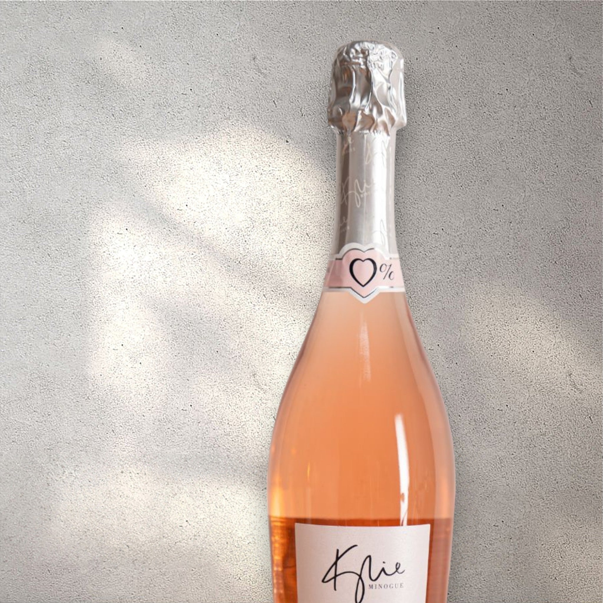 Closeup of a Kylie alcohol-free sparkling rosé wine bottle with a detailed label, set against a textured concrete wall, highlighting its refined design and blush pink colour.
