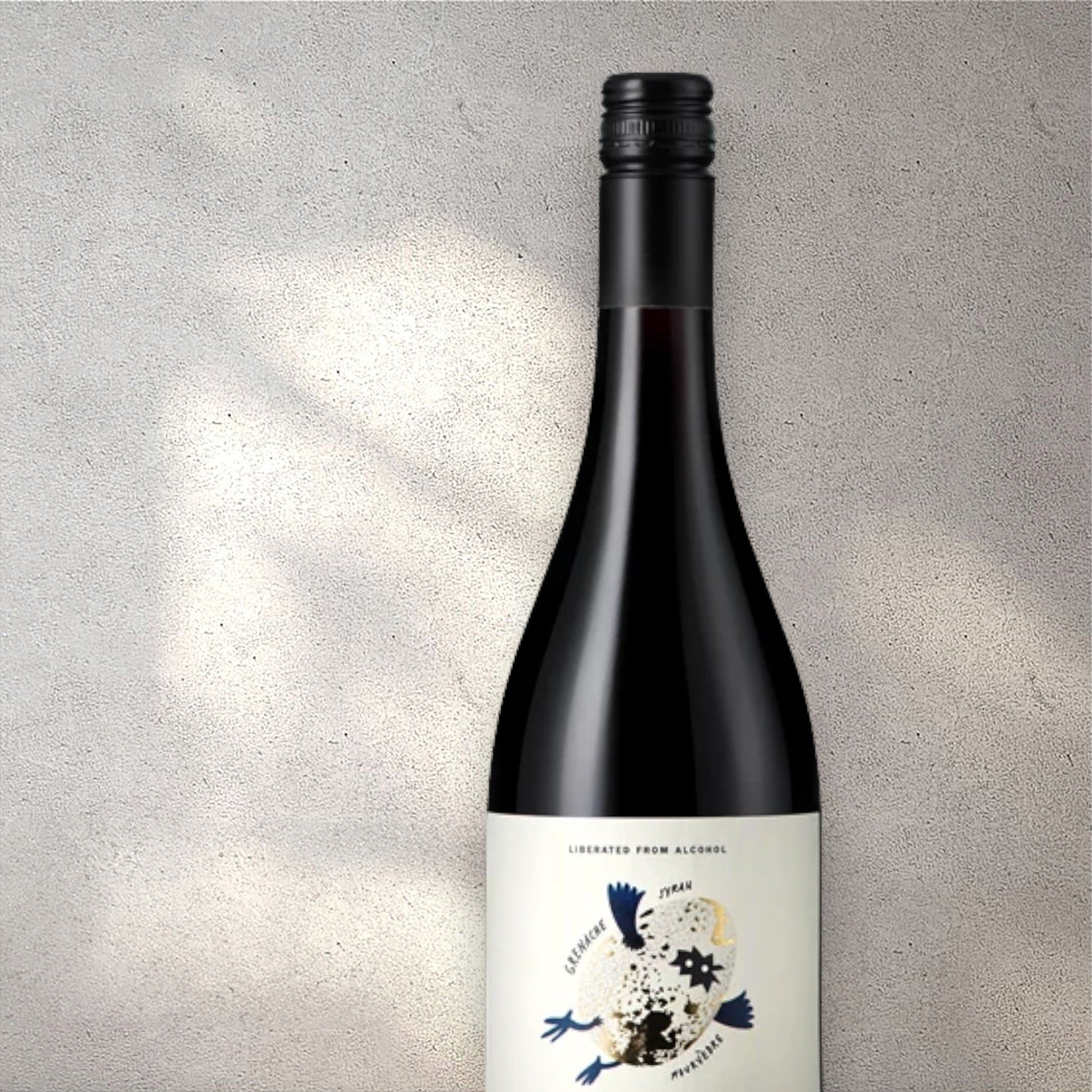 Closeup of an Oddbird Domaine GSM alcohol-free red wine bottle with a detailed label, set against a textured concrete wall, highlighting its refined design and deep red colour.