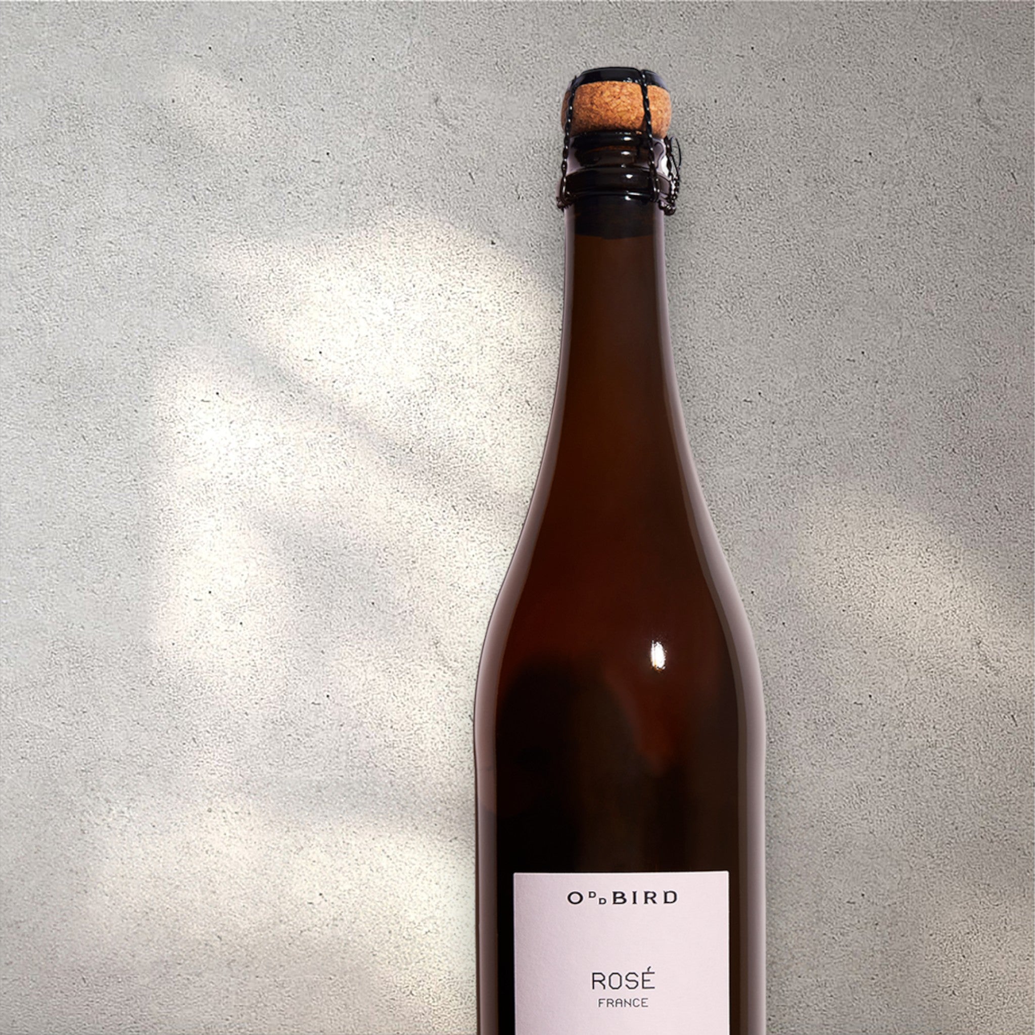 Closeup of an Oddbird alcohol-free sparkling rosé wine bottle with a detailed label, set against a textured concrete wall, highlighting its refined design and blush pink color.