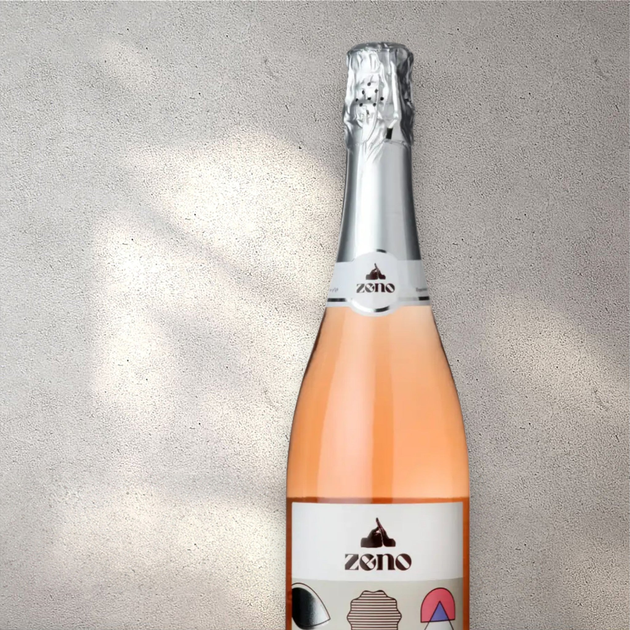 Closeup of a Zeno alcohol-free sparkling rosé wine bottle with a detailed label, set against a textured concrete wall, highlighting its refined design and blush pink color.